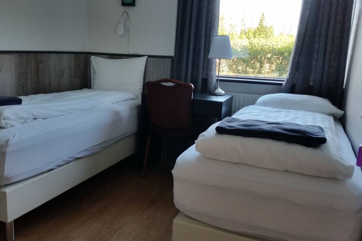 Quarto de hotel com duas camas de solteiro, uma escrivaninha, uma cadeira, um abajur e uma janela com cortinas.