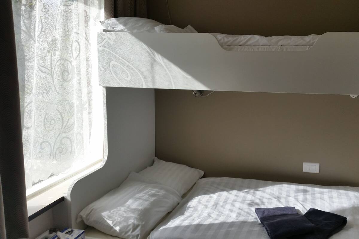 Uma cama beliche com uma janela à esquerda, uma mesa de cabeceira com abajur e uma tomada na parede.