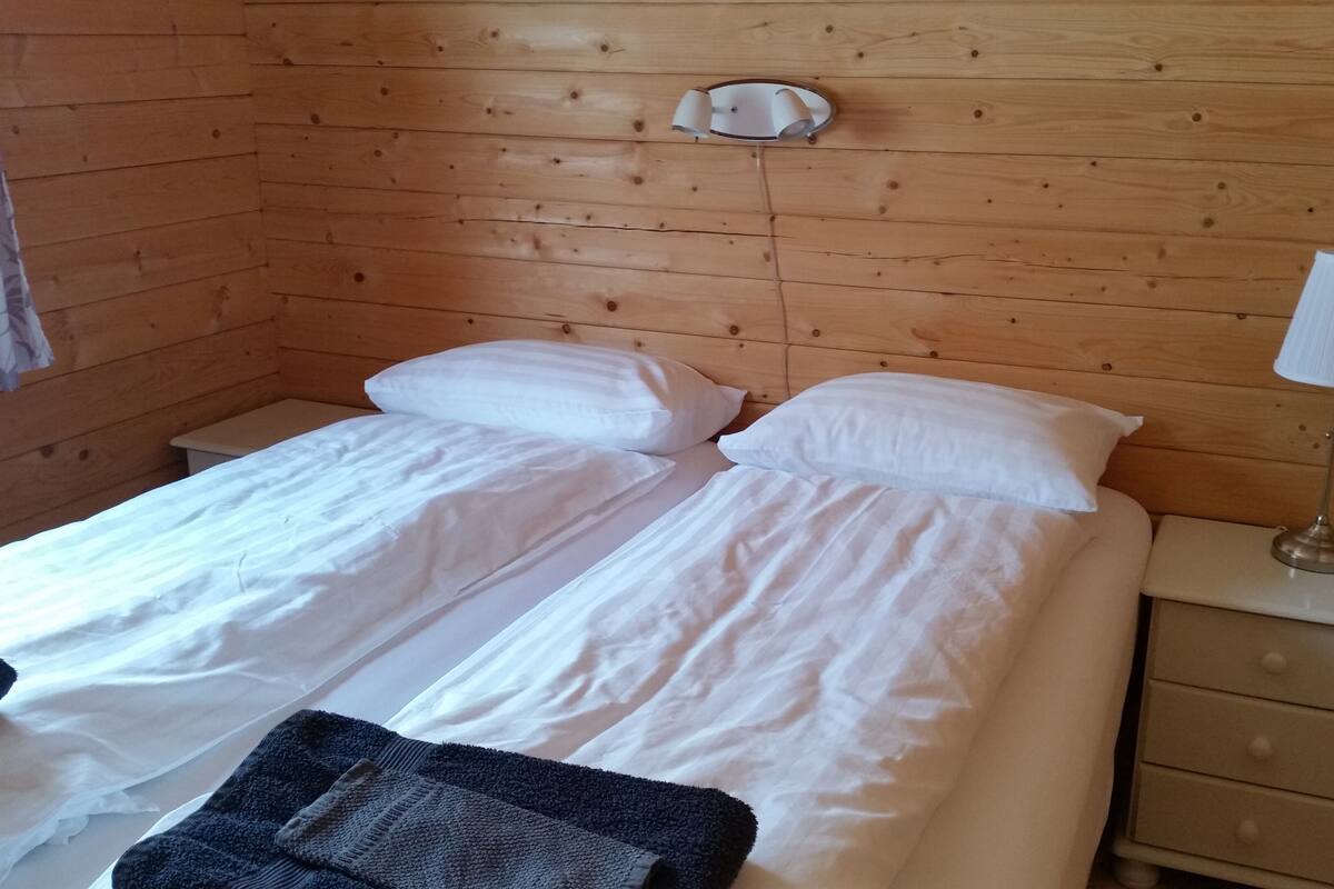 Quarto de cabana de madeira com duas camas, uma mesinha de cabeceira com abajur e cabeceira de madeira.