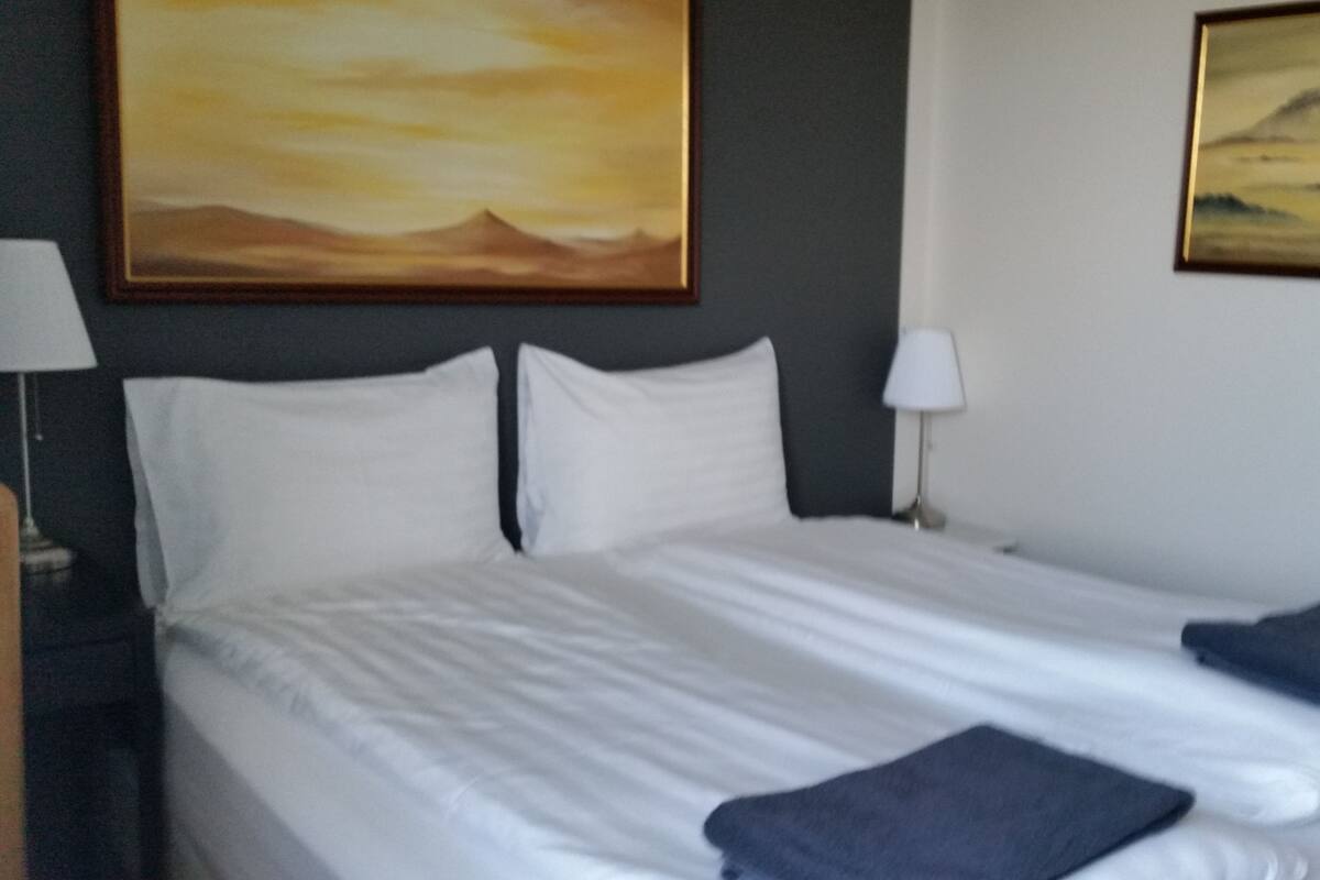 Quarto de hotel com uma cama grande, duas almofadas, mesa de cabeceira com abajur e duas pinturas na parede.