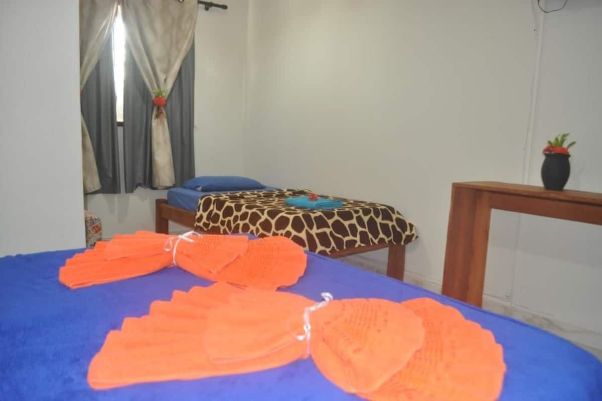 Ein Zimmer mit einem Bett, das mit einer blauen Decke und einer Bettwäsche mit Giraffenmuster bezogen ist, einem kleinen Tisch mit einem Topfblumen und Vorhängen mit einer Pflanzenbordüre.