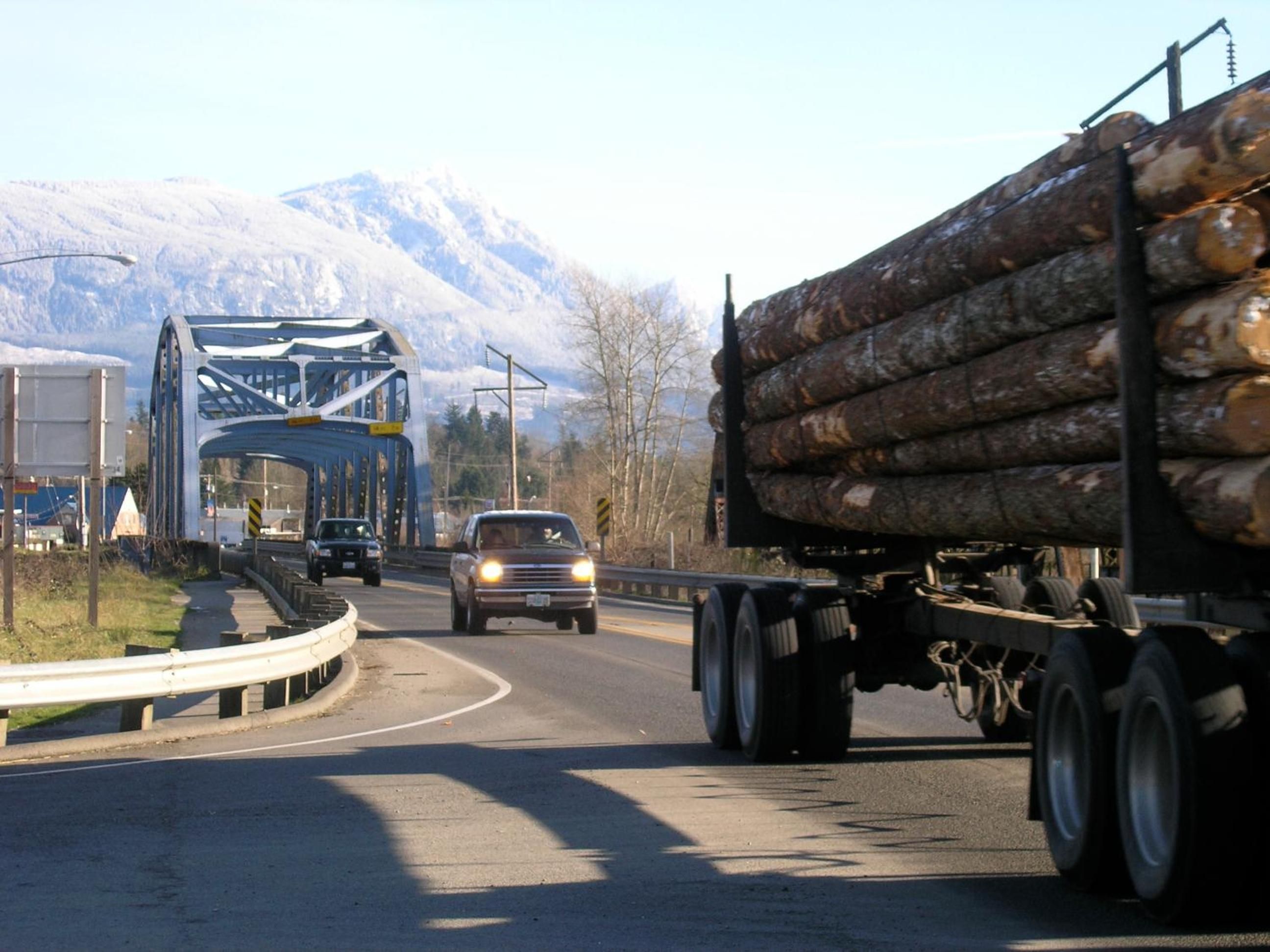 A highway with a large truck carrying logs and a blue bridge in the background.