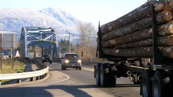 A highway with a large truck carrying logs and a blue bridge in the background.