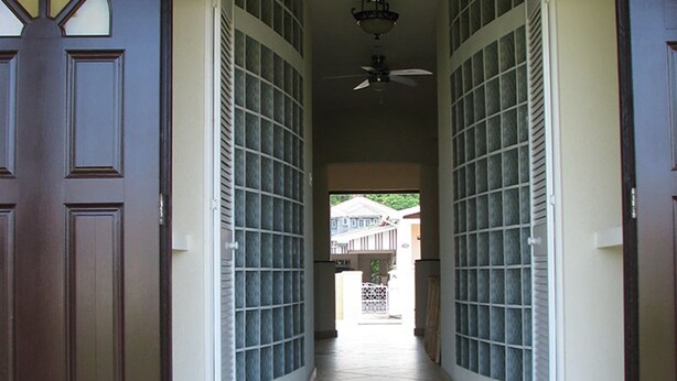 A hallway with a dark brown door on the left, a ceiling fan, and a view of a house in the distance. - Maya's Bajan Villas - Unit A (Oistins)