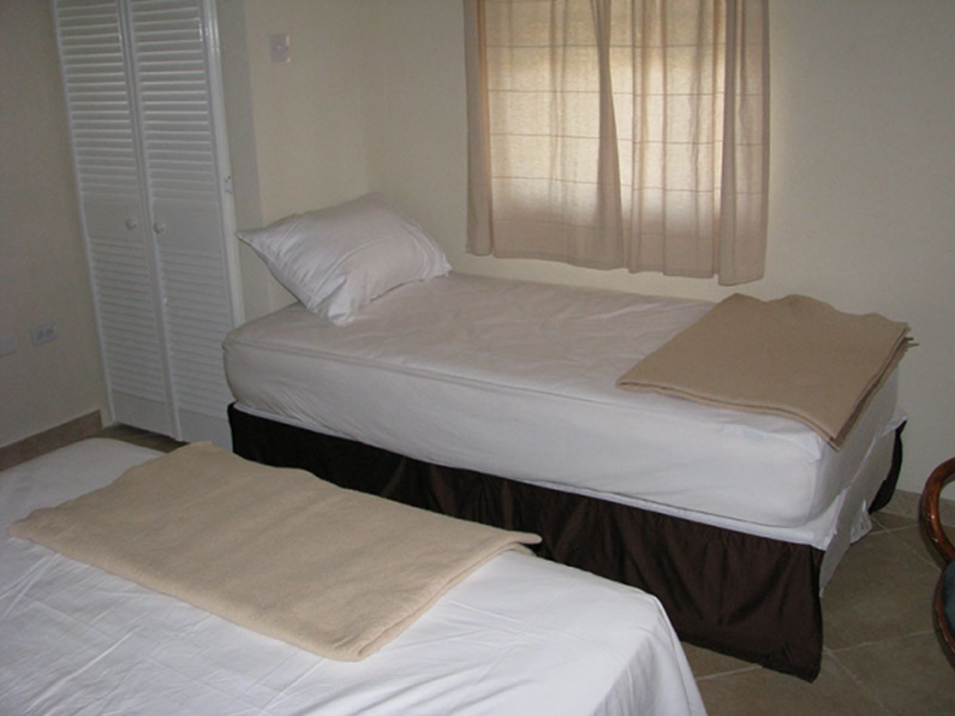 A small, single-bedroom room with two beds, a closet, and a window with sheer curtains.