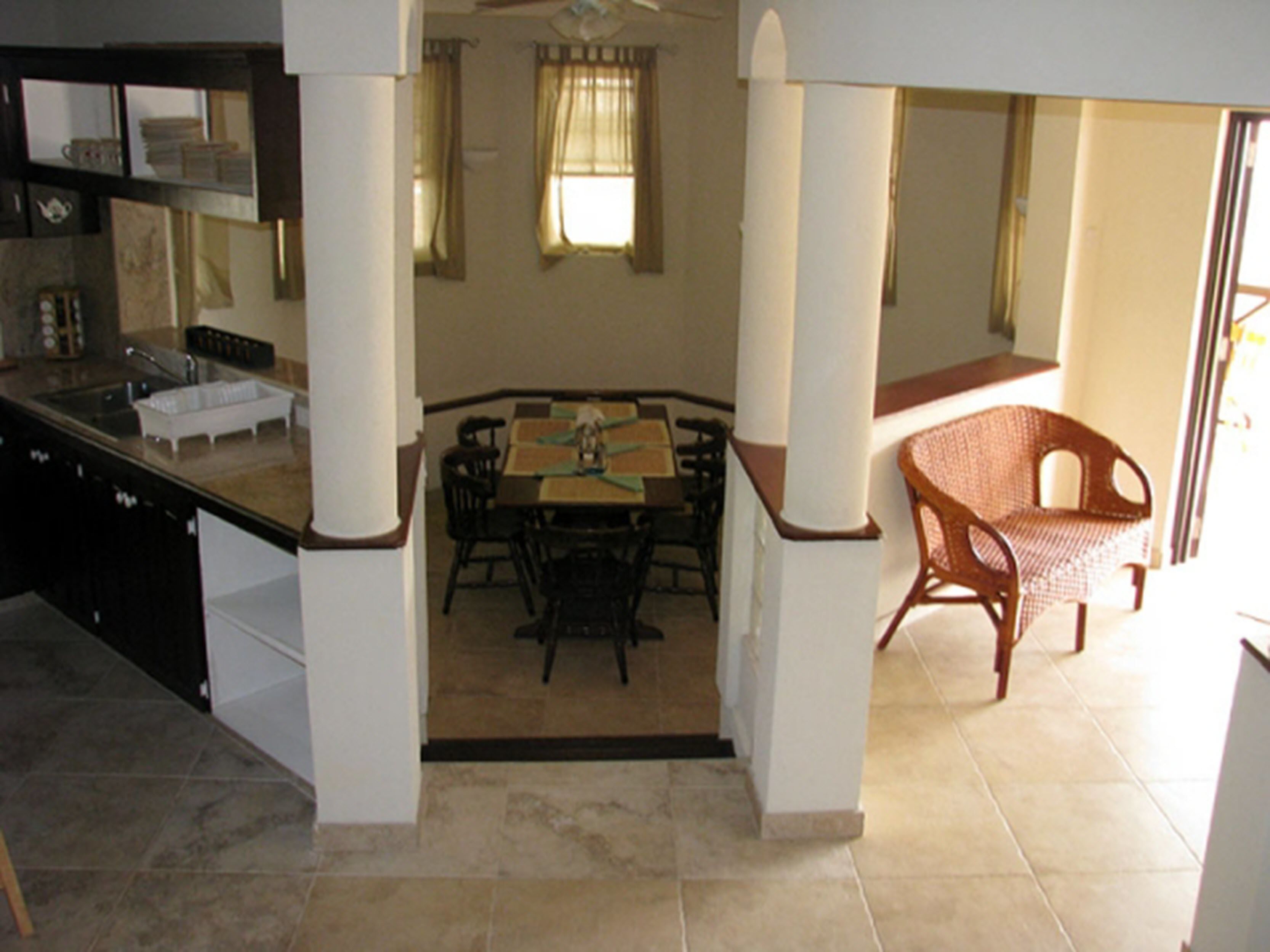 A dining area with a table and chairs, a kitchen with a sink and cabinets, and a wicker chair.