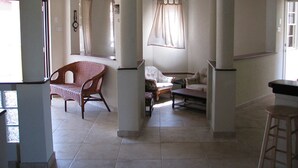 A spacious living area with a high ceiling, a ceiling fan, and a large window with curtains. There are several chairs and a sofa arranged for seating. - Maya's Bajan Villas - Unit A (Oistins)