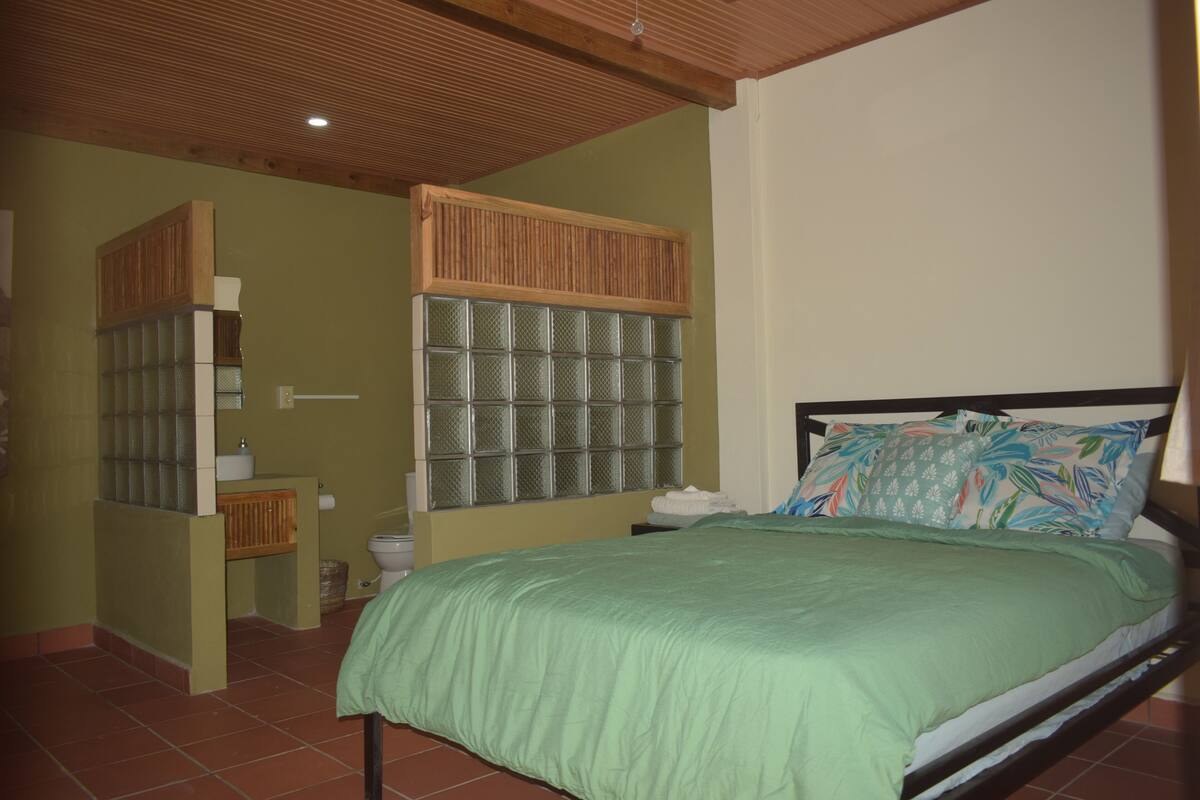 Un dormitorio con cama, cabecera de madera, colcha verde y baño con inodoro y lavabo.