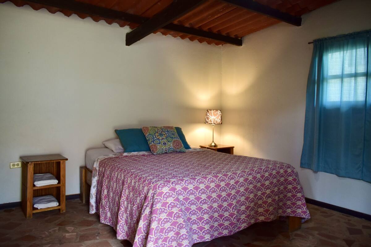 Un dormitorio con una cama, una mesita de noche, una lámpara y un estante con toallas.