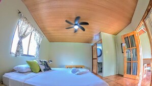 Una habitación con cama, ventilador de techo, ventana con cortinas y puerta que da a un baño.