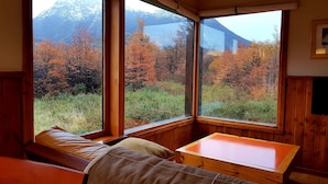 Habitación de esquina con paredes y banca de madera, y una mesa. Amplias ventanas ofrecen vistas a árboles otoñales y montañas.
