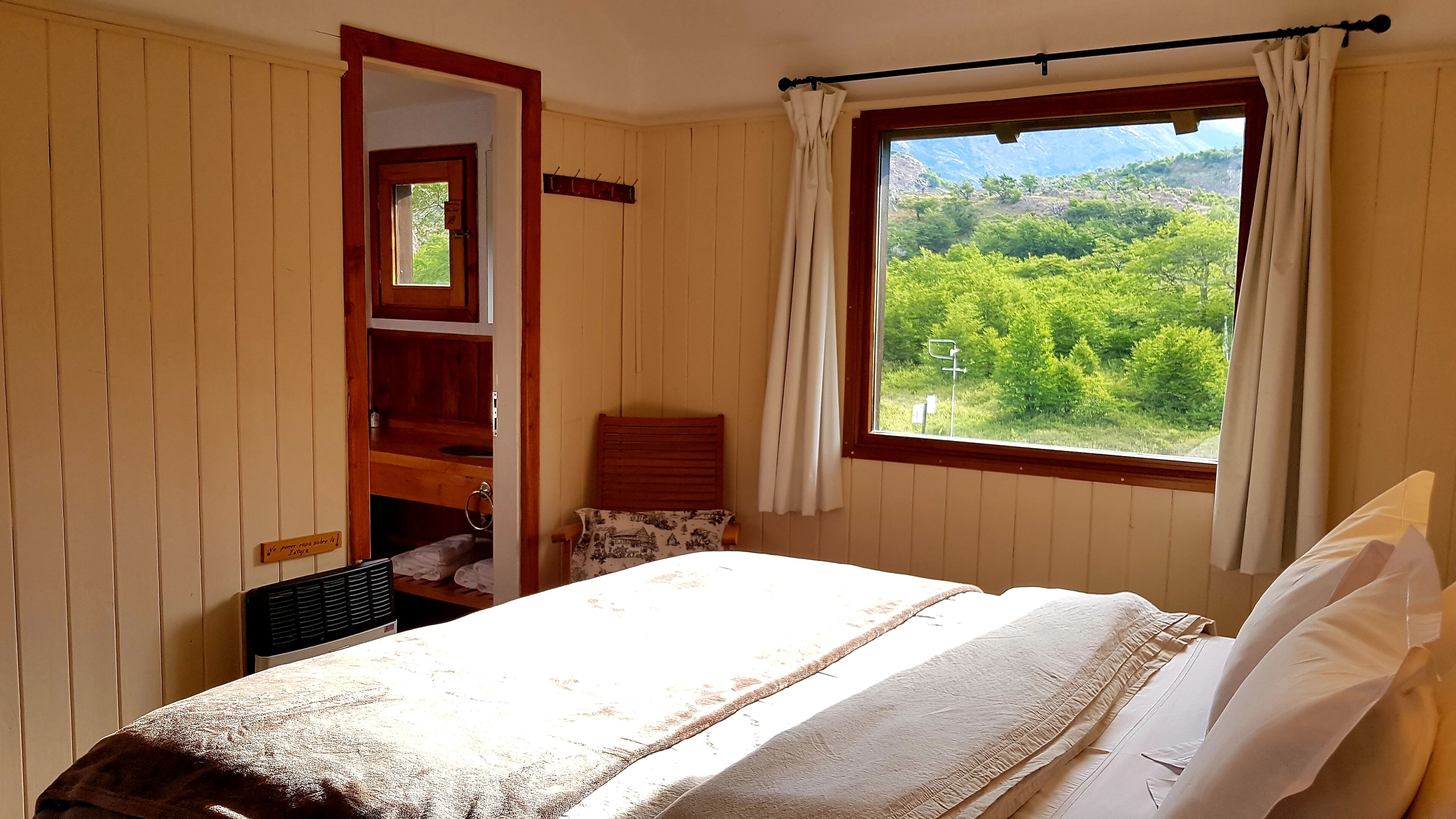 Une chambre à coucher avec un lit, une fenêtre avec des rideaux, une tête de lit en bois et une petite table avec une chaise.