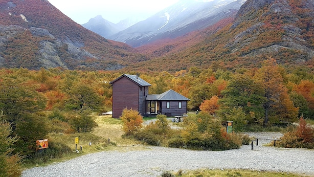 Una cabaña con un edificio principal grande y una estructura más pequeña adosada, rodeada de árboles de colores otoñales y montañas al fondo.