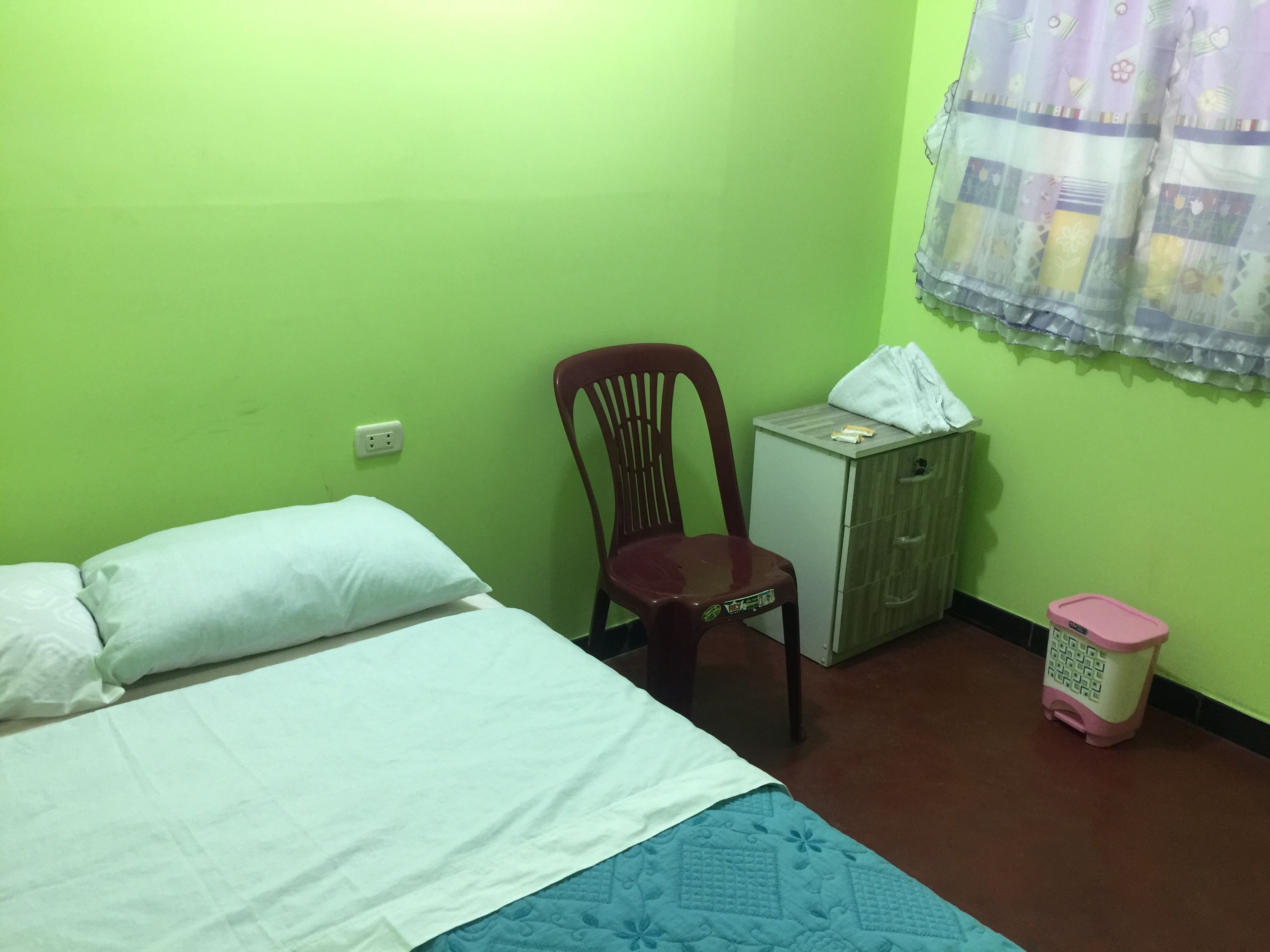 A small room with a bed, a chair, a small cabinet, and a trash bin.