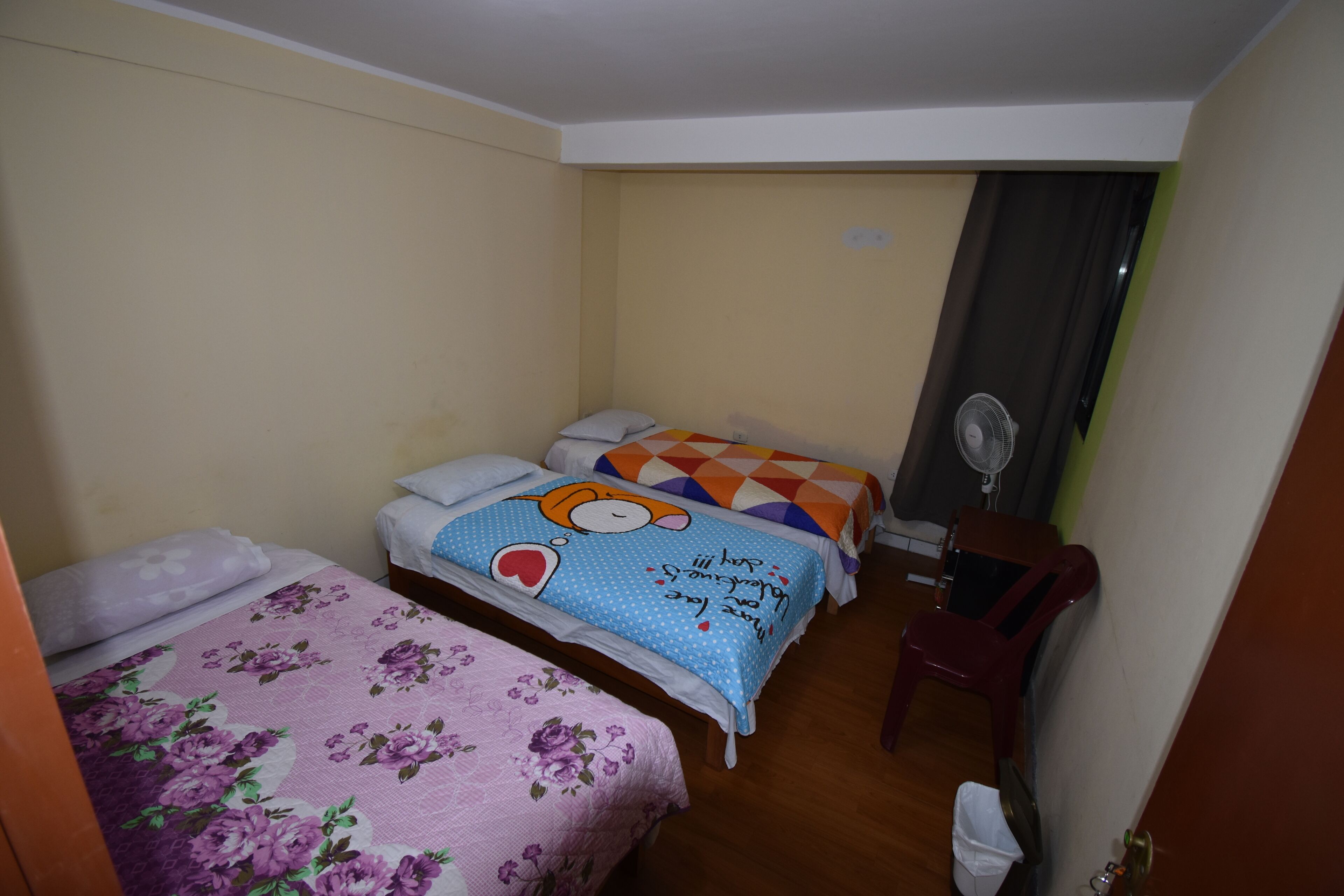 Habitación de hotel con dos camas, cada una con una colcha floral diferente, un escritorio de madera con una silla y un ventilador de pared.