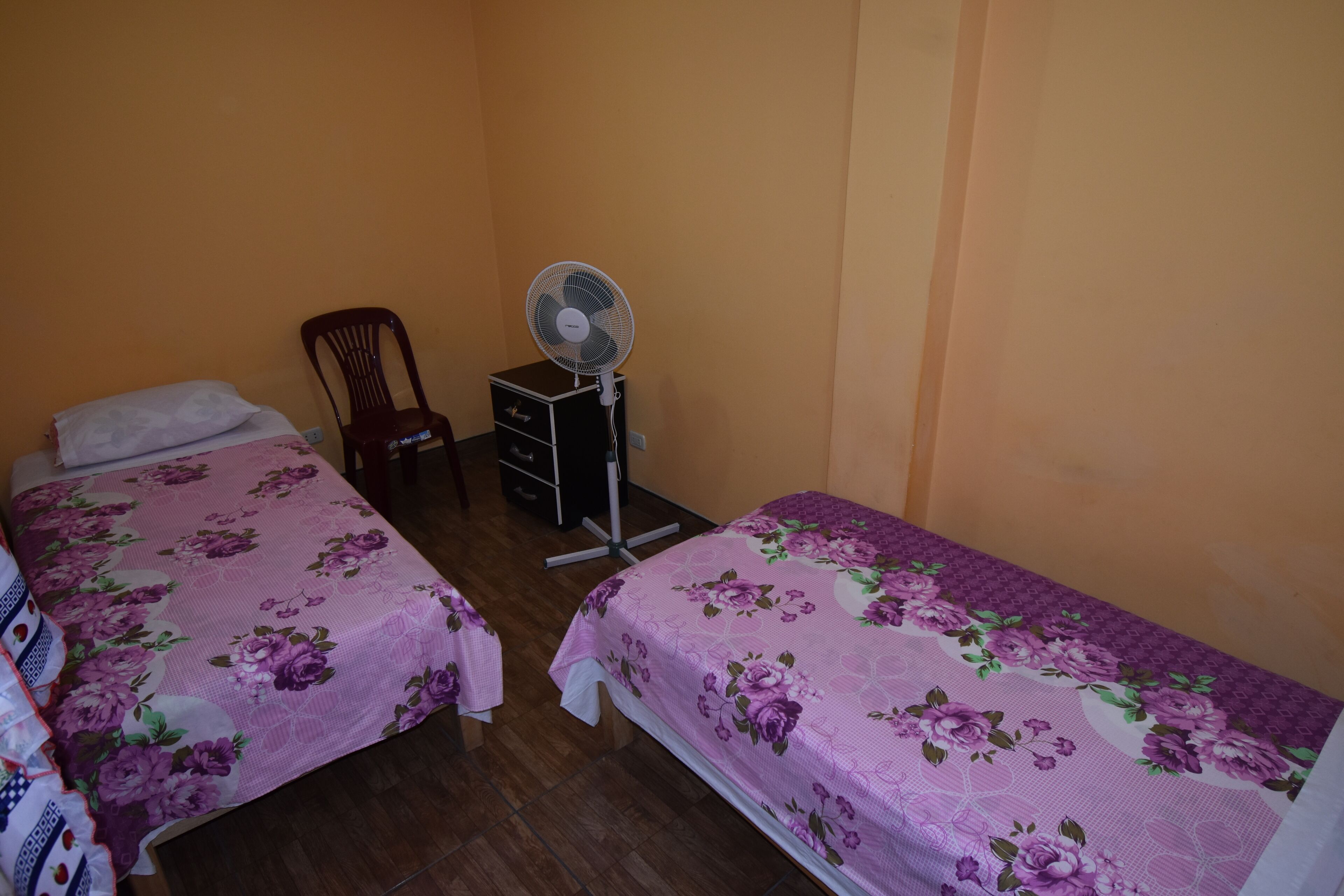 Habitación con dos camas, una silla, una mesita y un ventilador de pie.