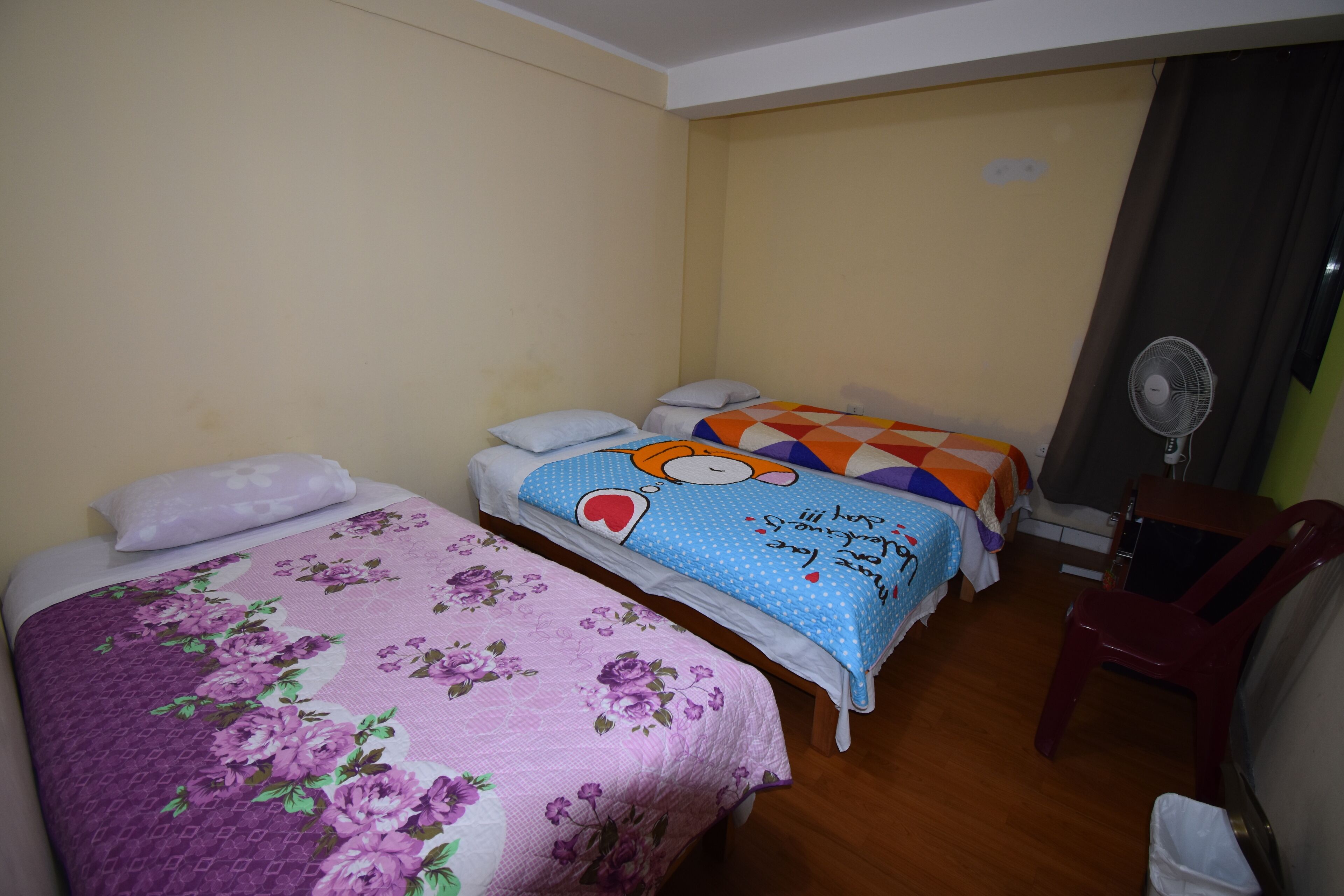 Habitación con dos camas, cada una con un edredón de estampado floral y geométrico. Hay una silla de madera y un ventilador de pie en la habitación.