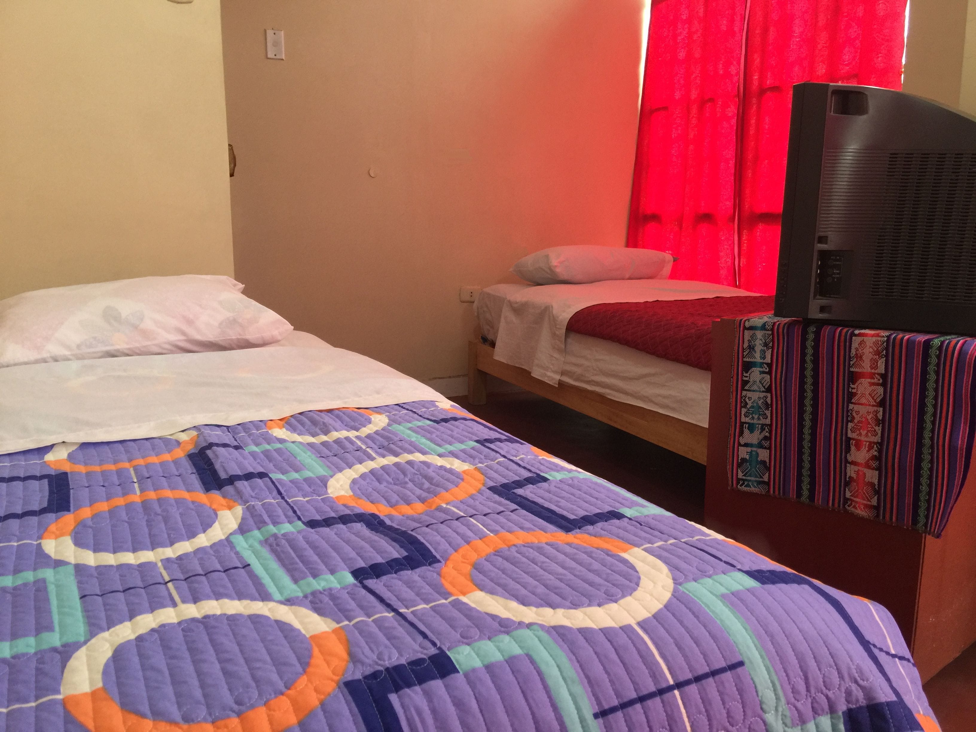 Una cama individual con un edredón de colores, una mesita de noche con un mantel estampado y un televisor sobre un mueble.