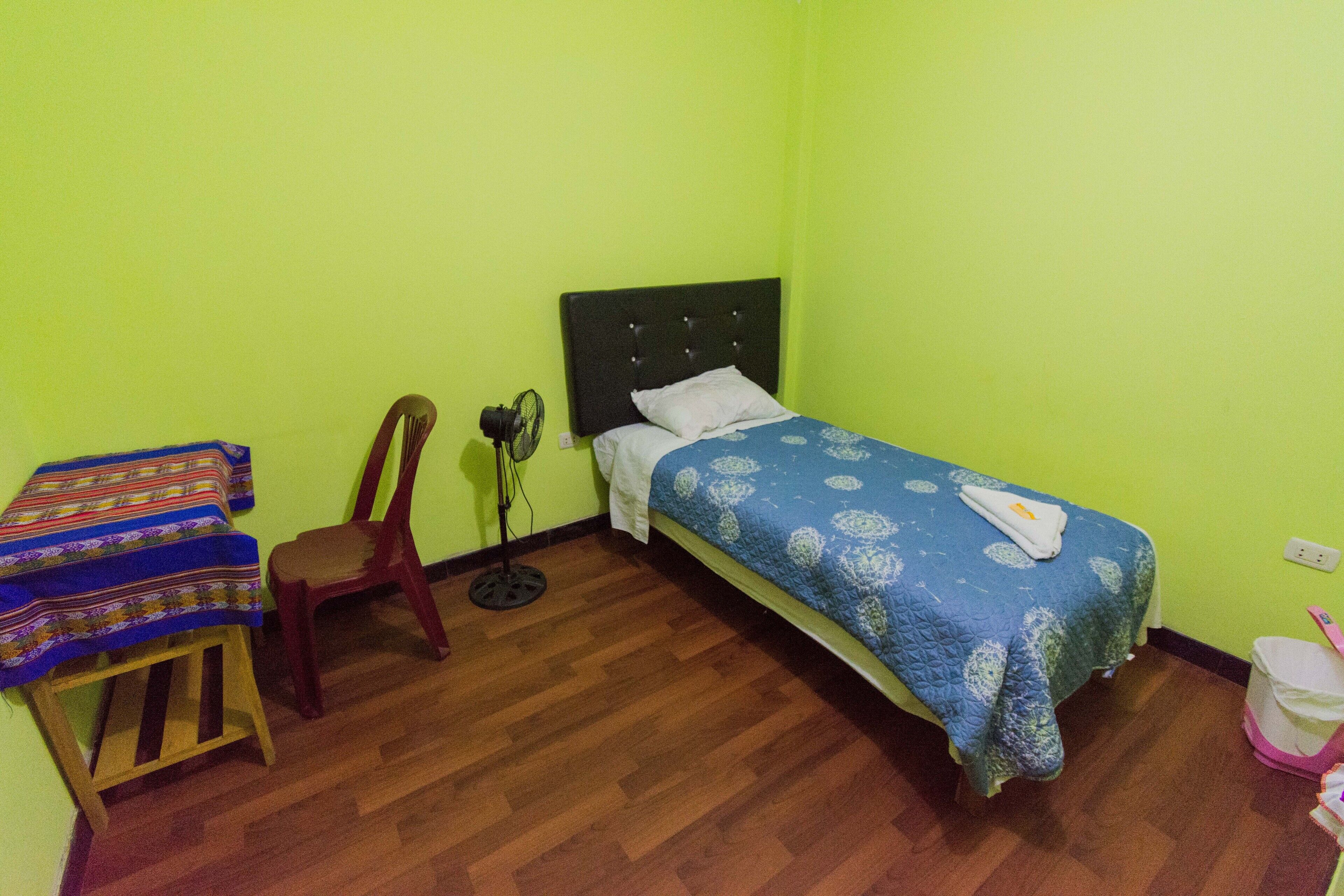 Ein kleines Zimmer mit einem Einzelbett, Holzboden, grüner Wand, einem Bett mit blauem, gemustertem Überwurf, einem Holzstuhl und einem kleinen Tisch mit farbigem Tischläufer.