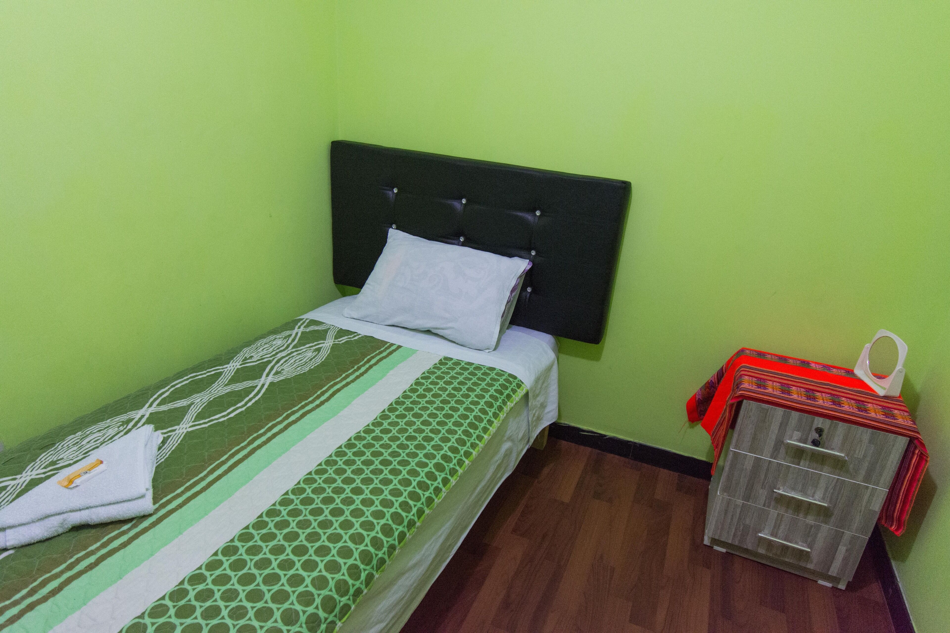 A single bed with a green and white patterned bedspread, a white pillow, and a wooden nightstand with drawers.