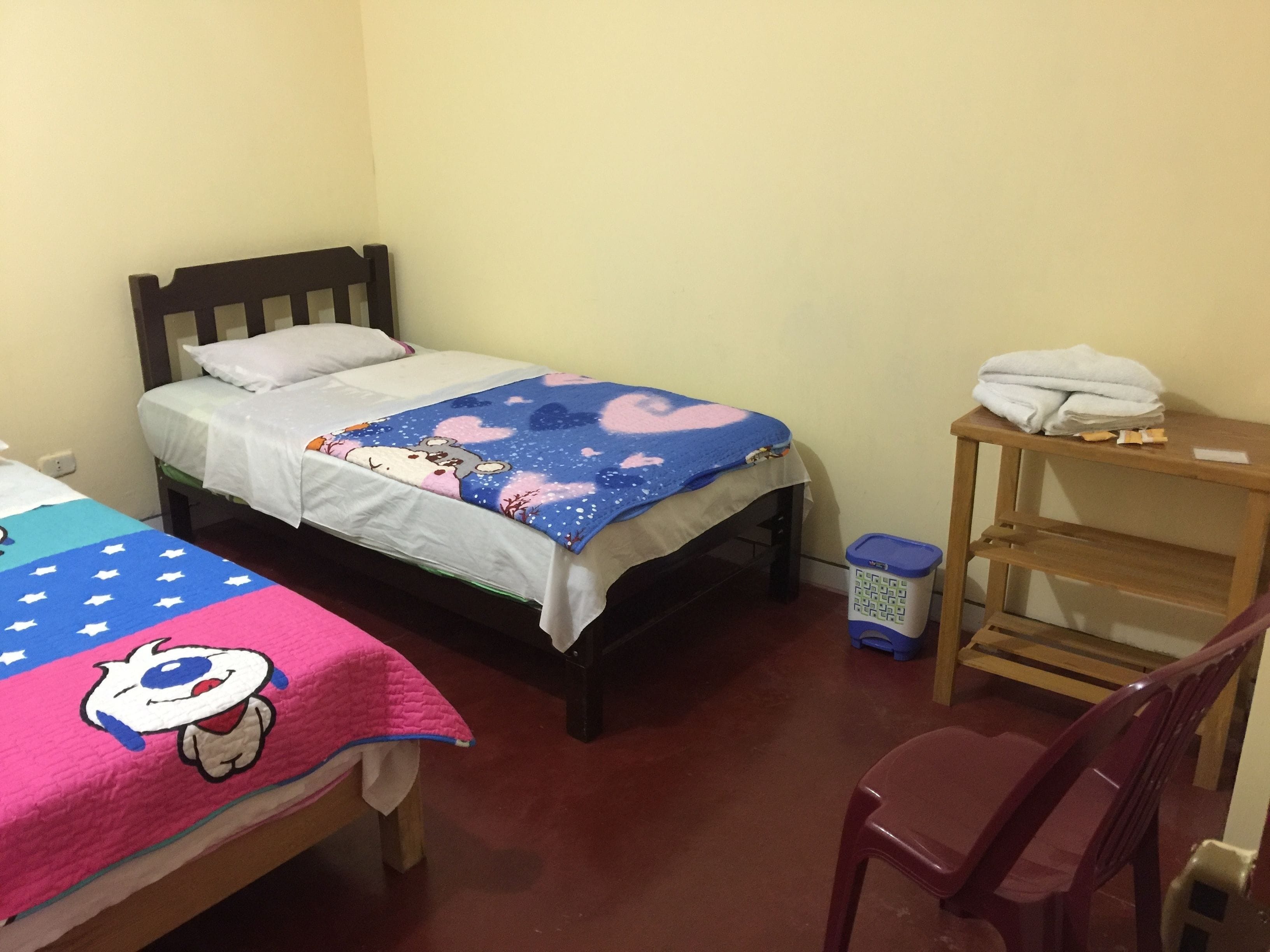 A small, simple room with two single beds, a wooden chair, a small table, and a trash bin.