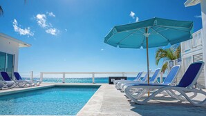 Ein Poolbereich mit Liegestühlen und einem großen Sonnenschirm, mit Blick auf einen klaren, blauen Himmel und das Meer.