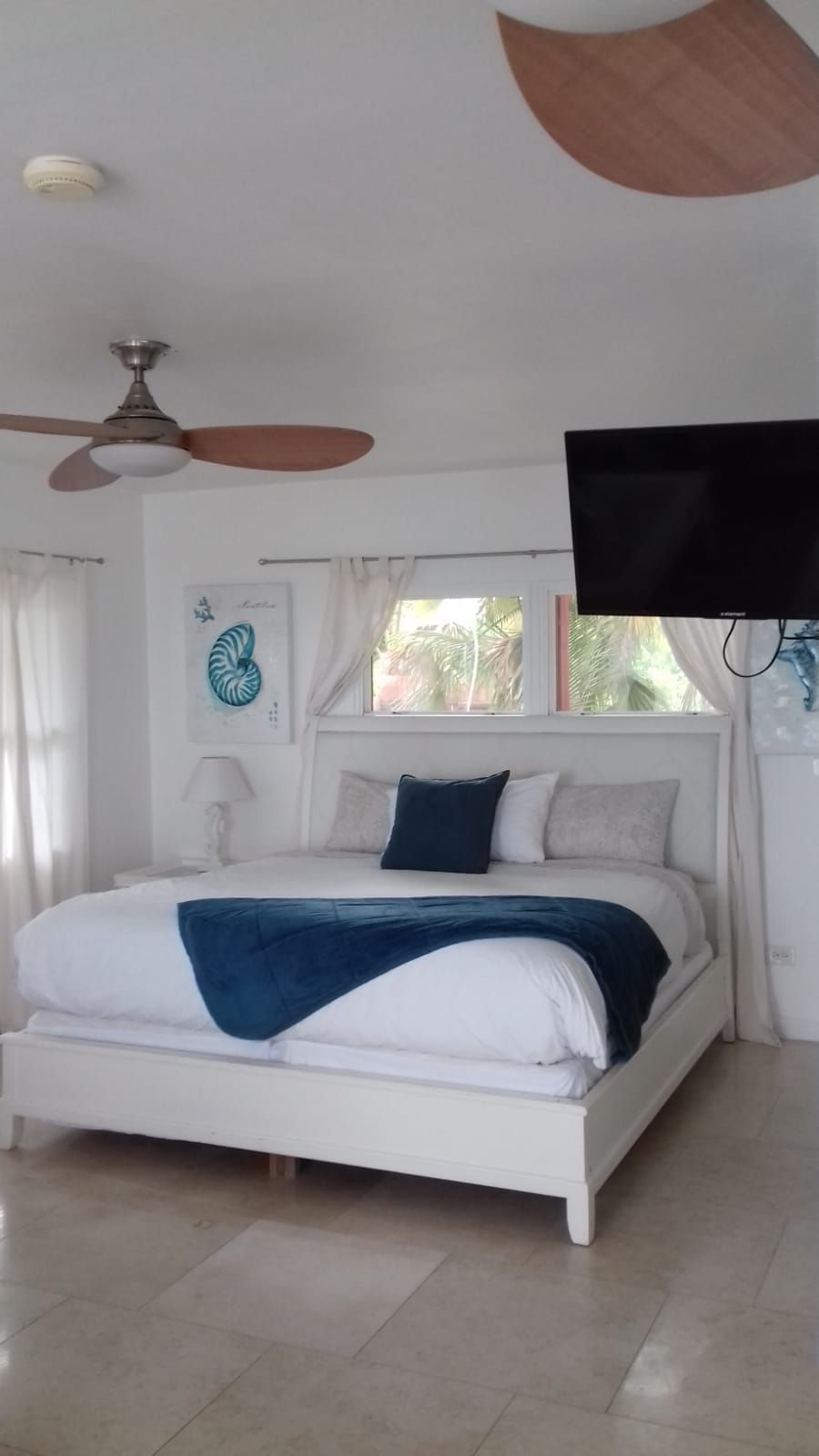 Ein Schlafzimmer mit einem Bett, einem Deckenventilator, einem an der Wand montierten Fernseher und einem Fenster mit Vorhängen.