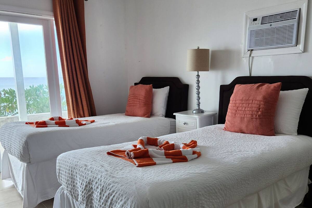 A hotel room with two beds, a window with curtains, a bedside lamp, and a view of the sea.