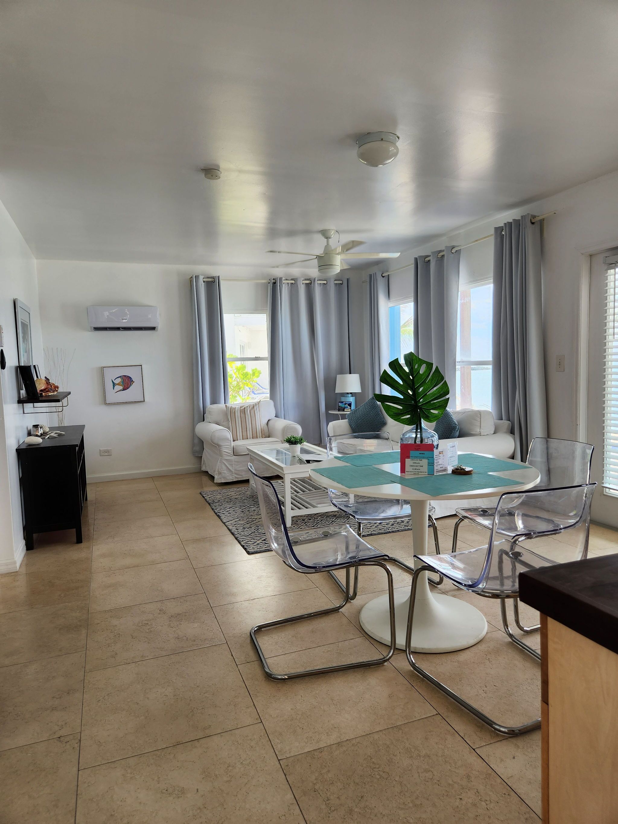 Un salon moderne comprenant un coin repas, des meubles blancs, un ventilateur de plafond et une grande fenêtre avec des rideaux.