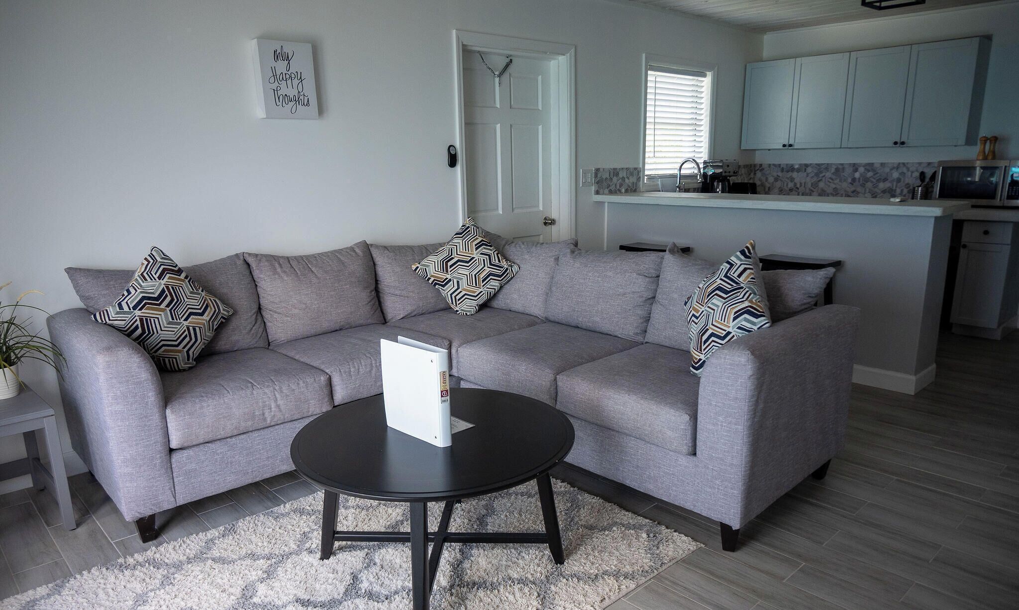 A living room with a grey sofa, black coffee table, patterned cushions, a white door, and a kitchen in the background.