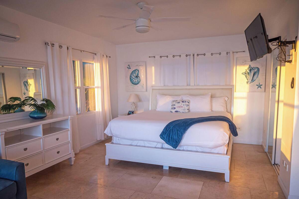 A bedroom with a bed, a dresser, a mirror, a ceiling fan, and a TV mounted on the wall.