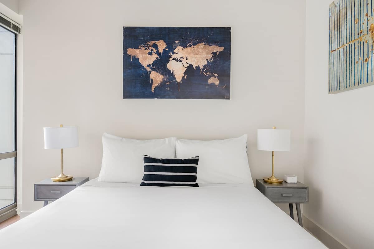 A neatly made bed with white linens, two bedside lamps, a world map artwork on the wall, and a small bedside table with a lamp and a charging device.
