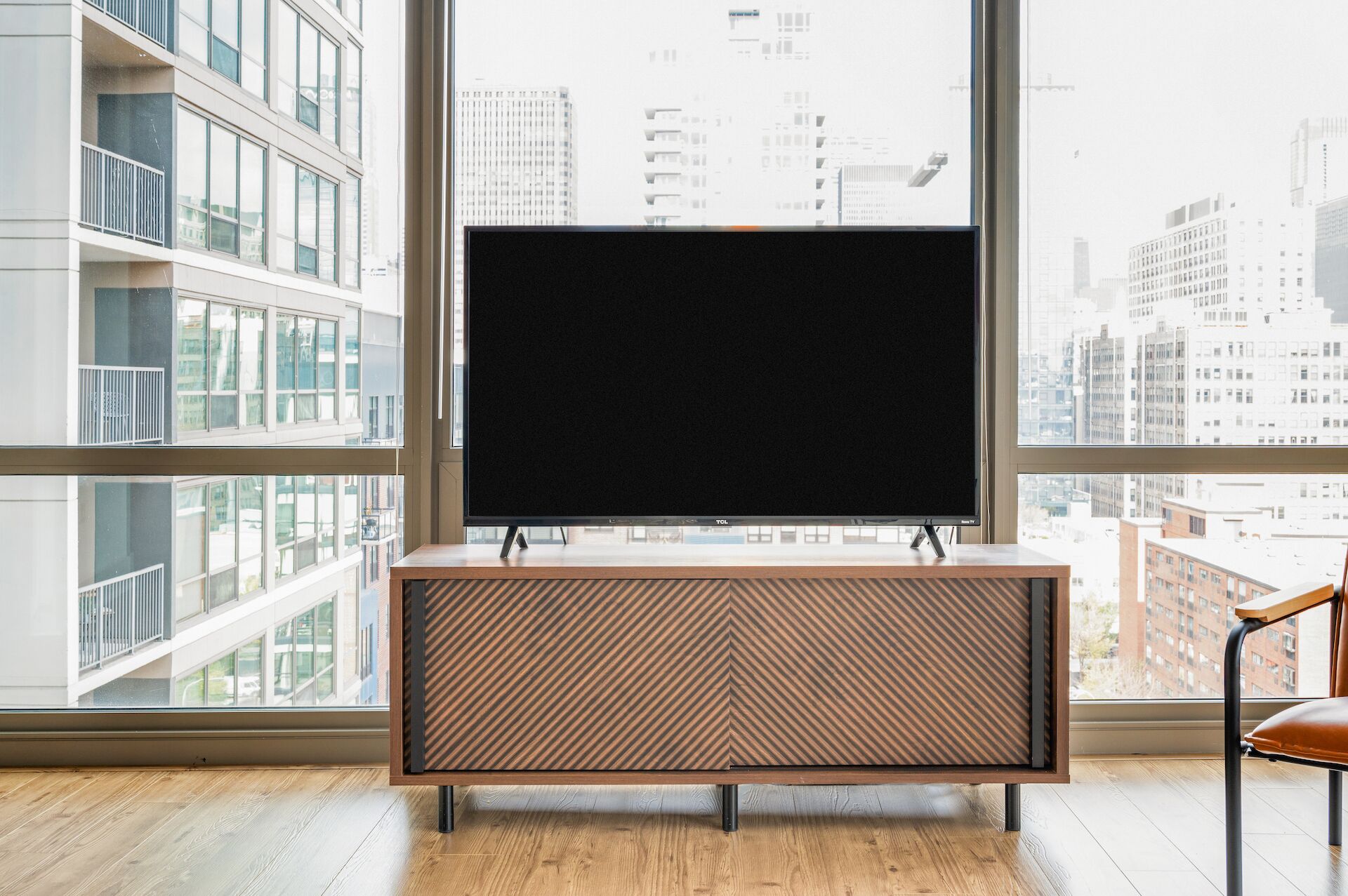 A modern living room with a large flat-screen TV on a wooden console table, large windows offering a city view, and a chair in the foreground.