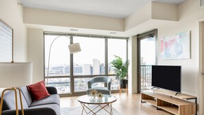 Een moderne woonkamer met een grijze bank, een ronde salontafel, een televisie op een houten dressoir en een groot raam met uitzicht op de stad.