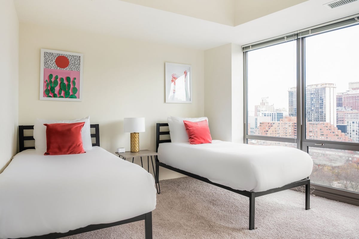 A hotel room with two beds, a nightstand, and a large window offering a city view.