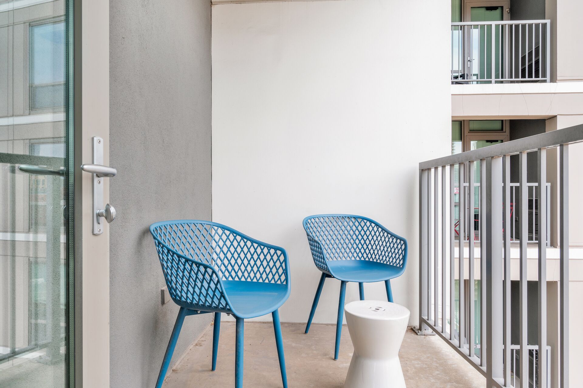 A balcony with two blue chairs and a white table, a glass door, and a white railing.
