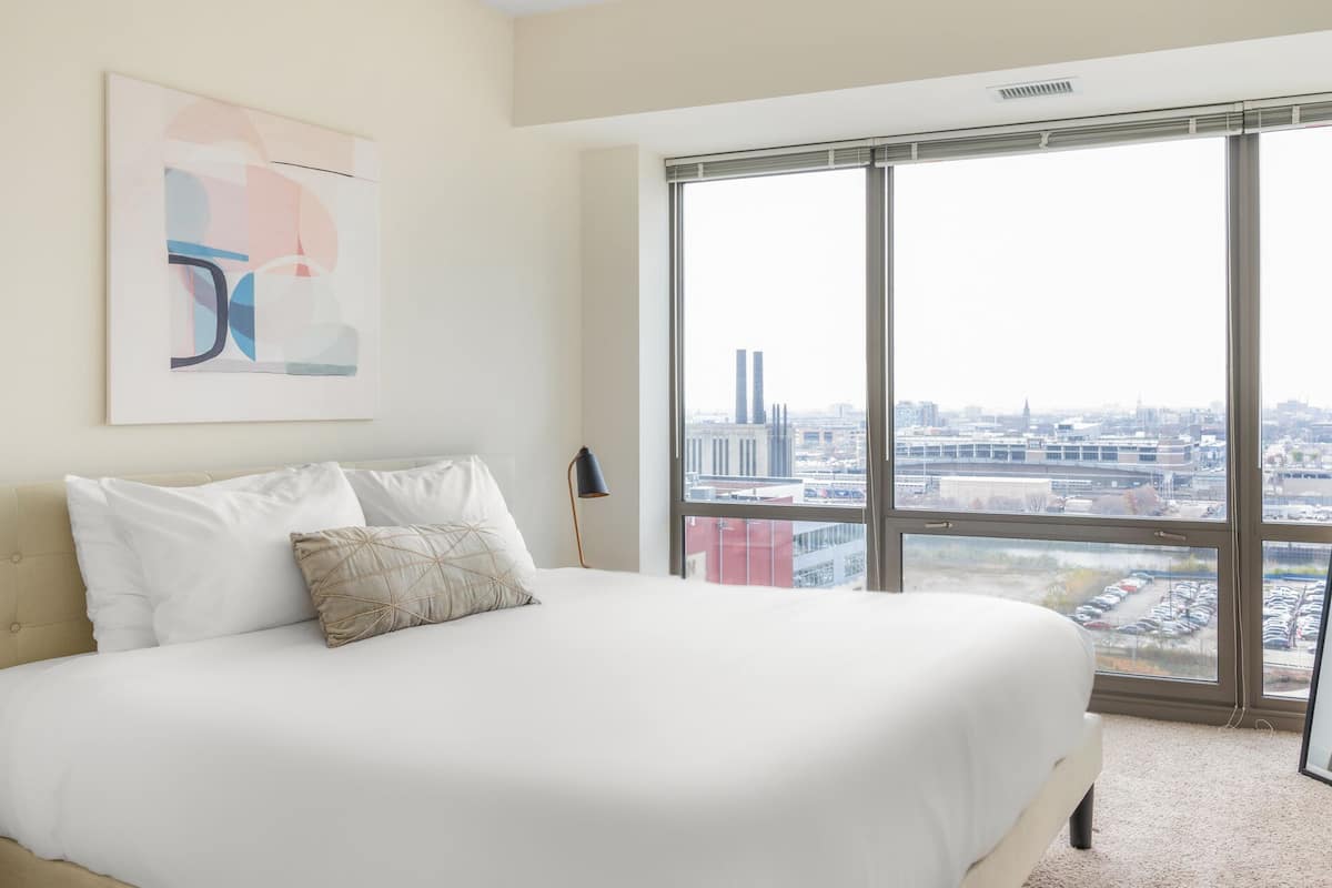 A bedroom with a large bed, a city view through floor-to-ceiling windows, and a piece of abstract art on the wall.