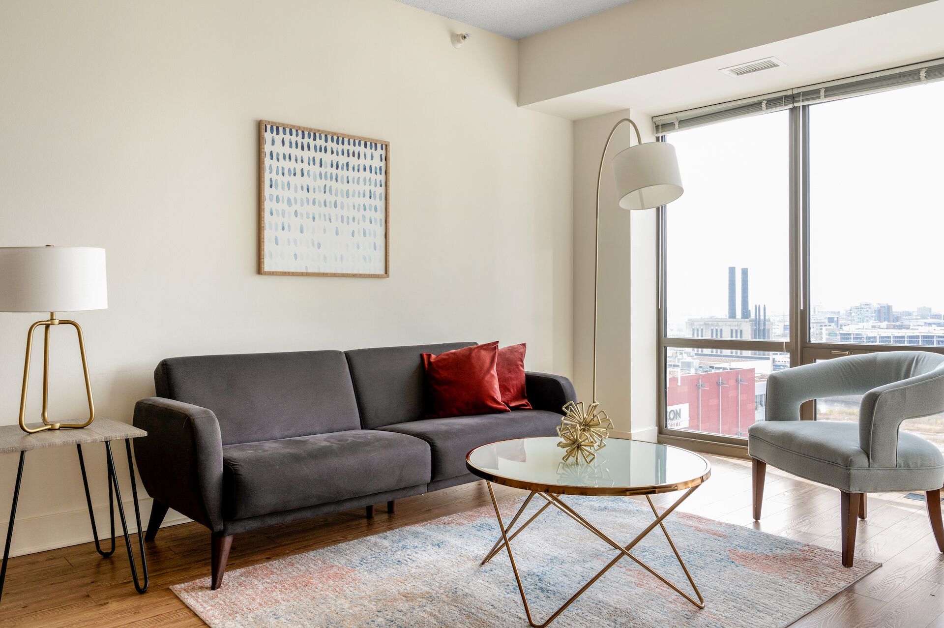 Una sala de estar moderna con un sofá gris, una mesa de centro redonda de vidrio y un sillón gris. Un ventanal ofrece vistas a la ciudad.