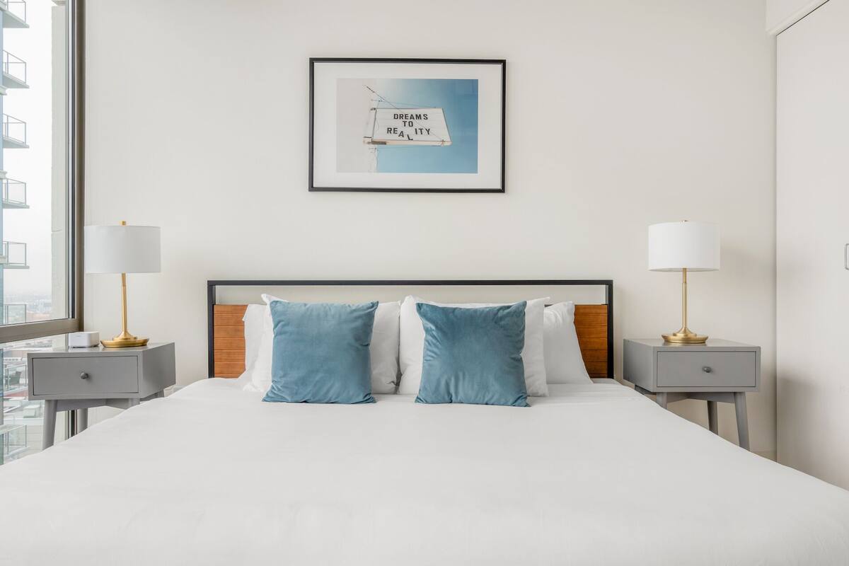 A hotel room with a large bed, two bedside tables with lamps, a framed picture on the wall, and a view of the city through the window.