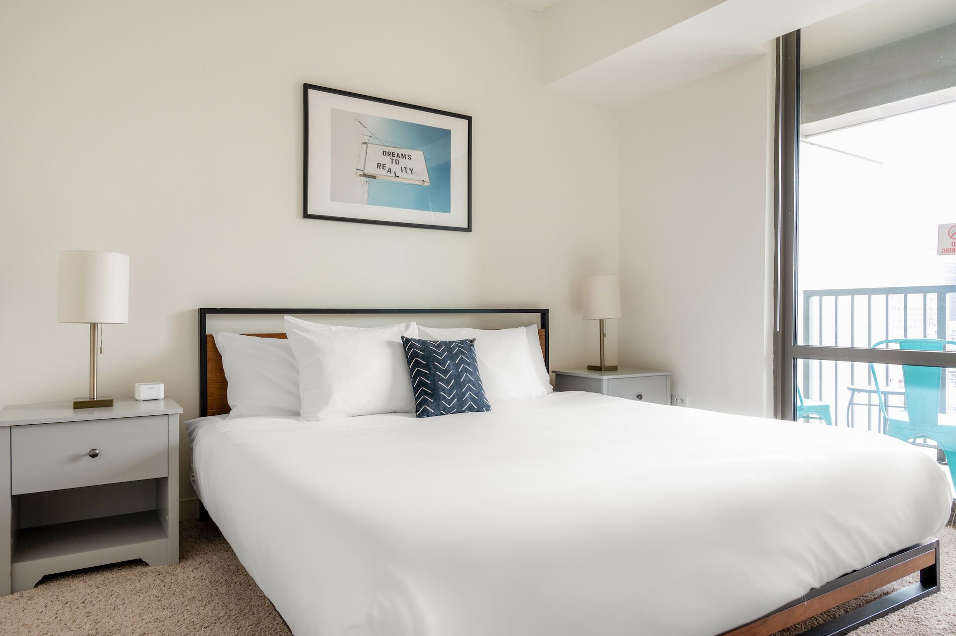 Quarto de hotel bem arrumado com uma cama grande, duas mesas de cabeceira, uma frase inspiradora emoldurada na parede e vista para a varanda.