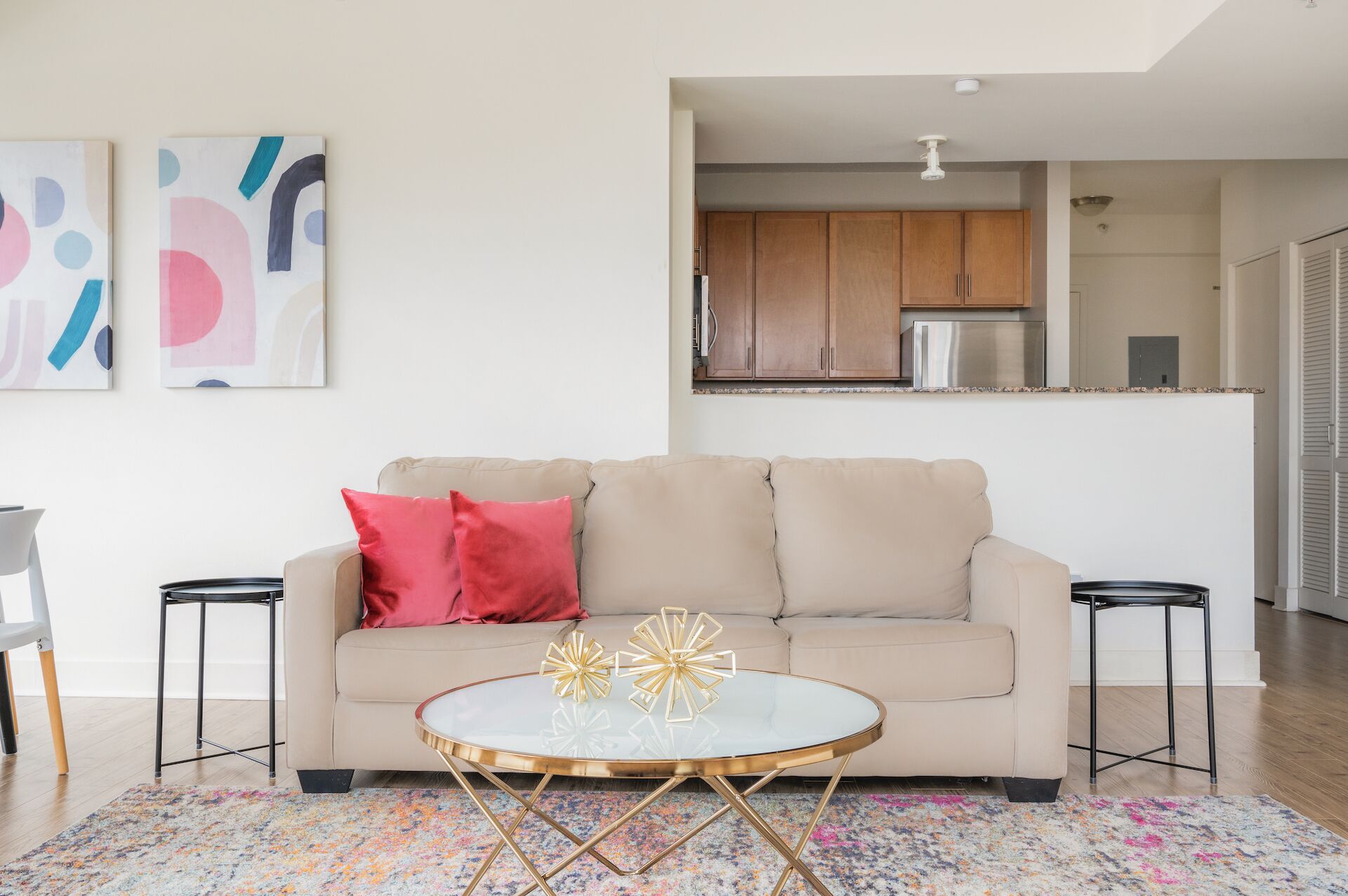 Ein modernes Wohnzimmer mit einem beigefarbenen Sofa, roten Kissen und einem Glastisch mit goldenen Beinen. An den Wänden hängen abstrakte Gemälde und im Hintergrund ist eine Küchenzeile zu sehen.