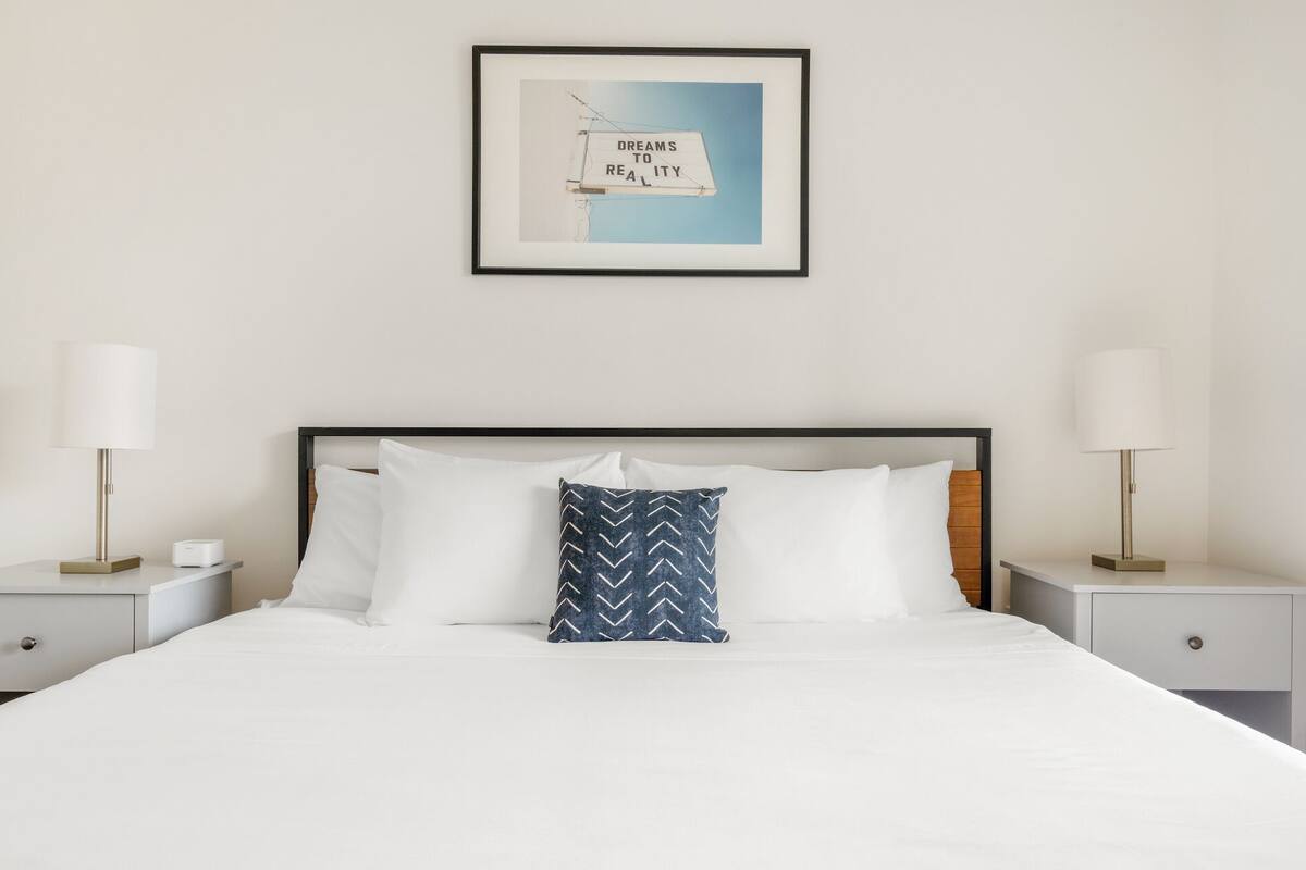A neatly made bed with white linens, a blue patterned pillow, and a framed picture above it.