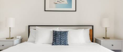 A neatly made bed with white linens, a blue patterned pillow, and a framed picture above it.
