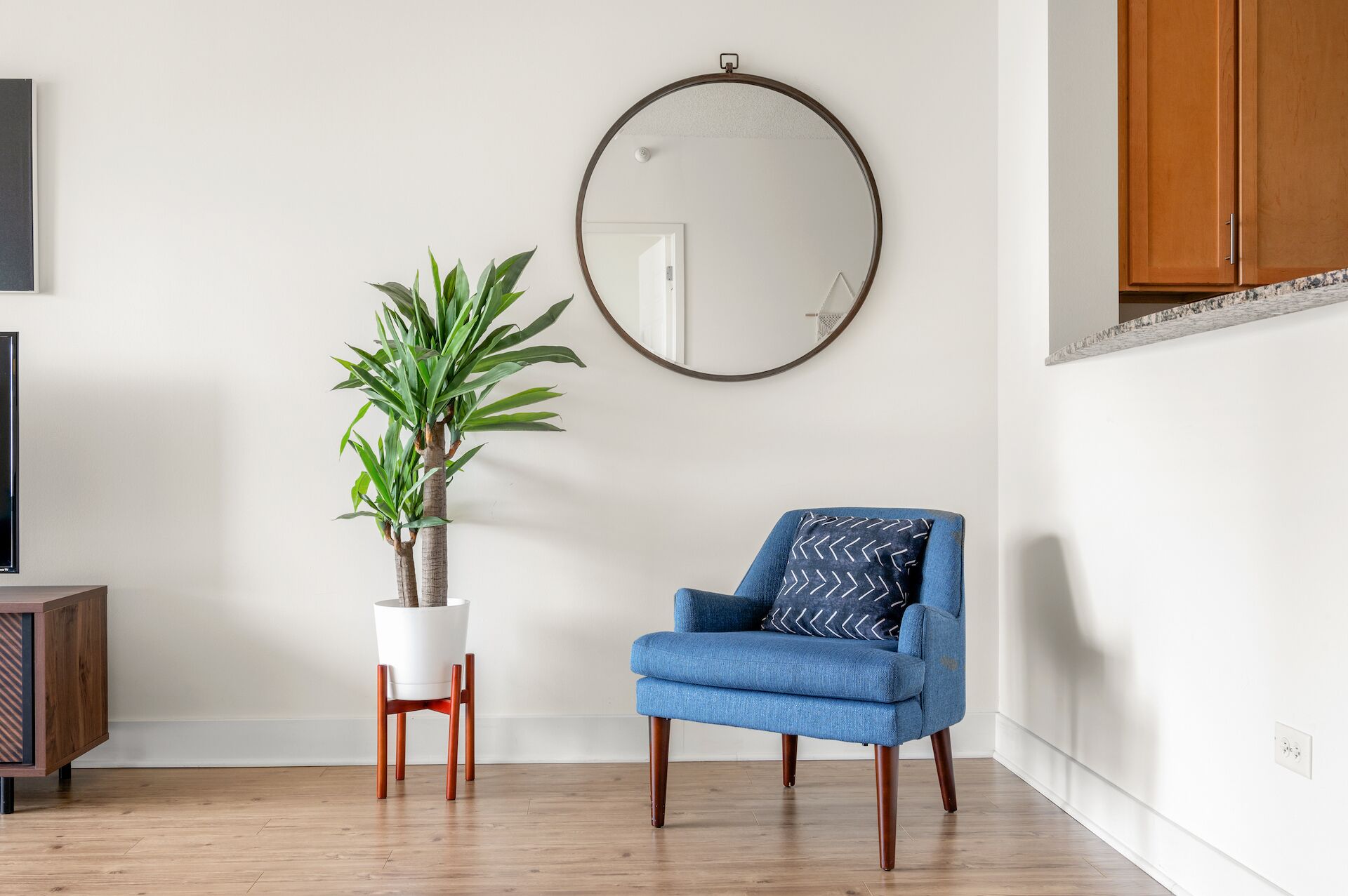 A room with a blue armchair, a potted plant, a round mirror, and a wooden cabinet.