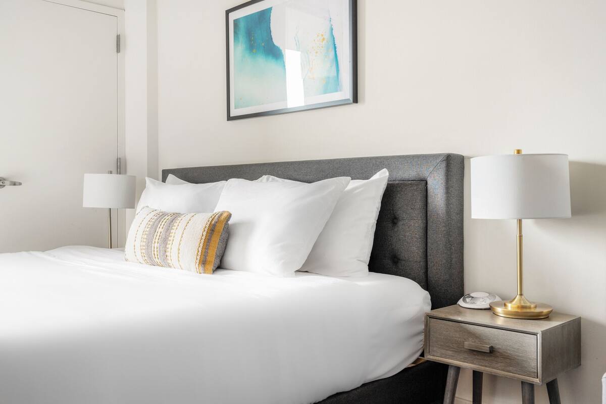 A neatly made bed with white linens and a grey headboard, flanked by two bedside tables with lamps and a bedside clock.