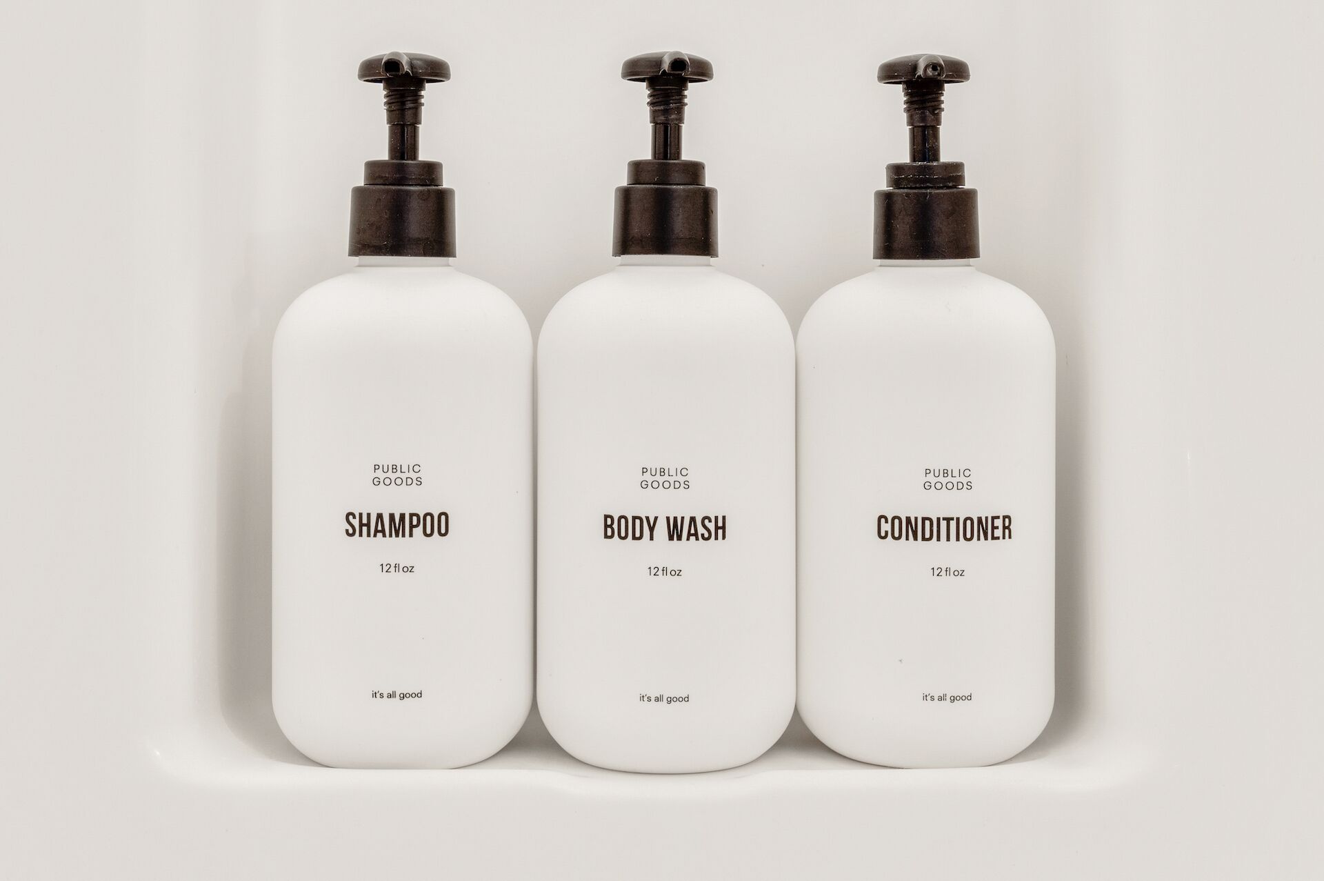 Tre vita flaskor med produkter från Public Goods: schampo, duschkräm och balsam, var och en märkt med en volym på 12 flytande uns och frasen ”allt är bra”.