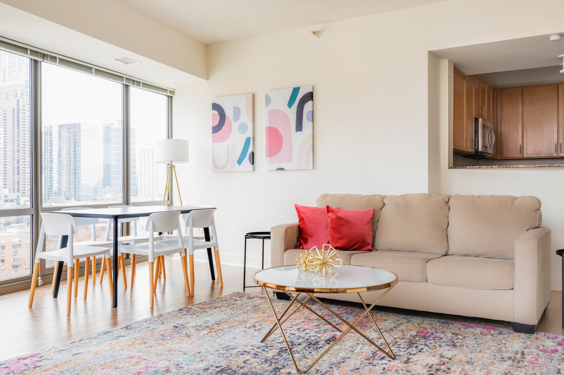 A modern living room with a sofa, a coffee table, and a dining area with chairs. There are abstract paintings on the wall and a view of buildings through large windows.