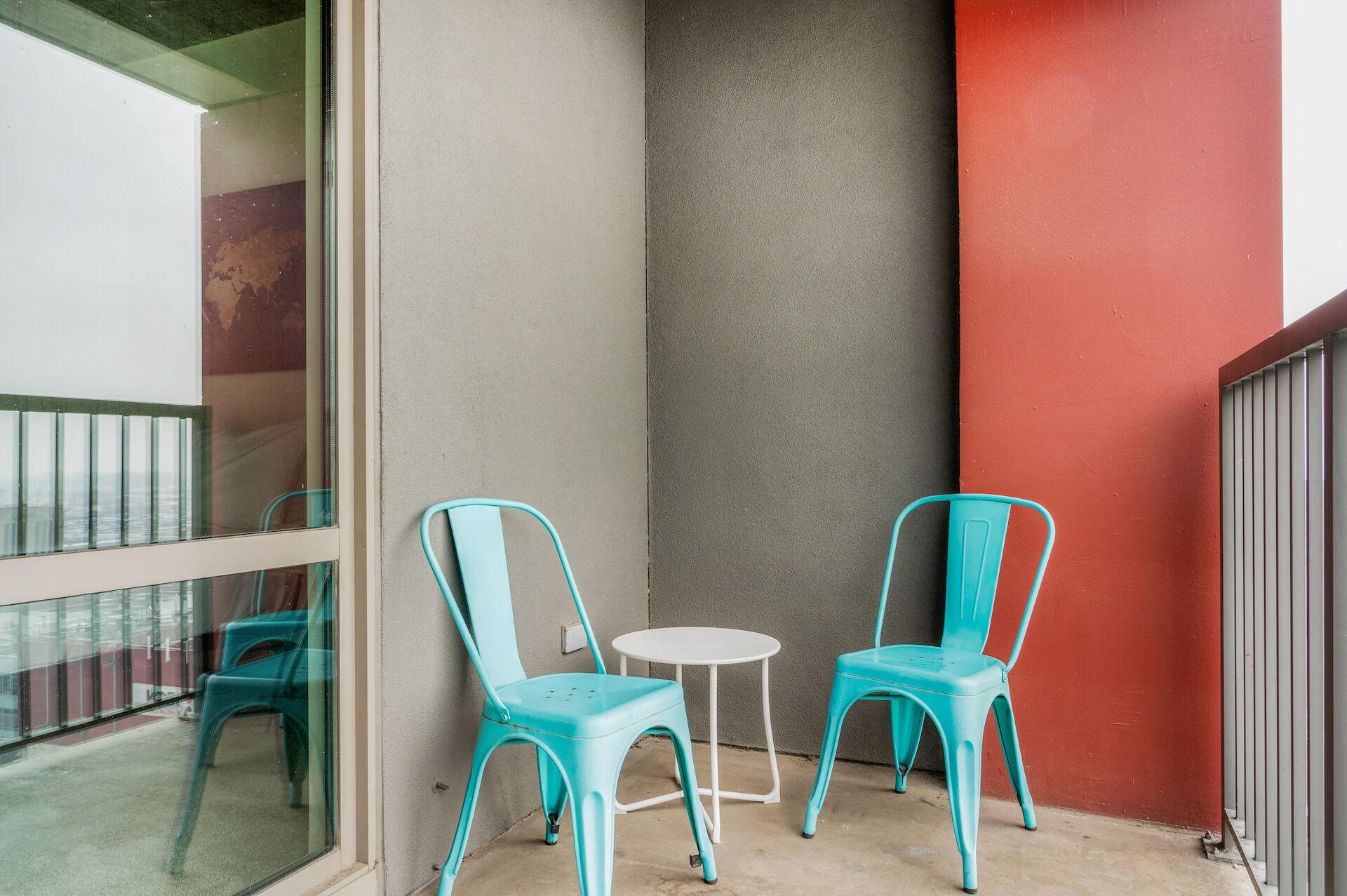 Un pequeño espacio al aire libre con dos sillas turquesas y una mesa blanca, junto a un edificio con una pared roja y un pilar gris.