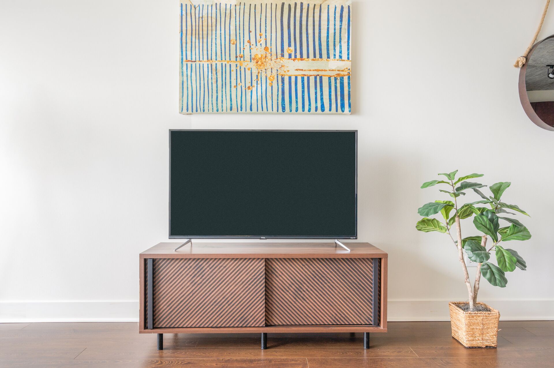 Un téléviseur à écran plat posé sur un meuble en bois orné de lignes diagonales, un panier tressé contenant une plante et, au mur, un tableau abstrait aux couleurs bleu et blanc.