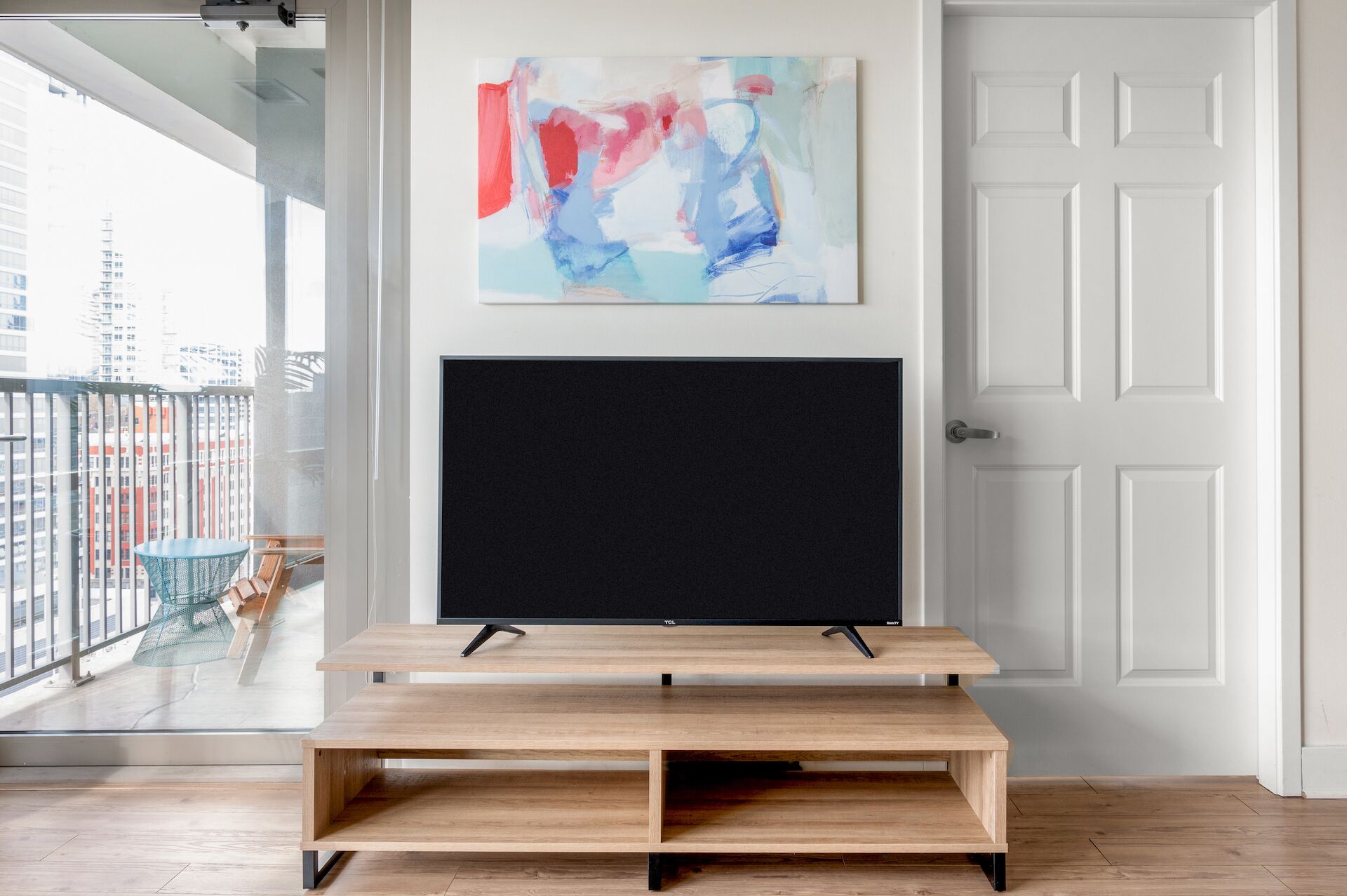 Un salon moderne avec un grand téléviseur à écran plat posé sur un meuble en bois, un balcon offrant une vue sur les immeubles et une porte blanche.
